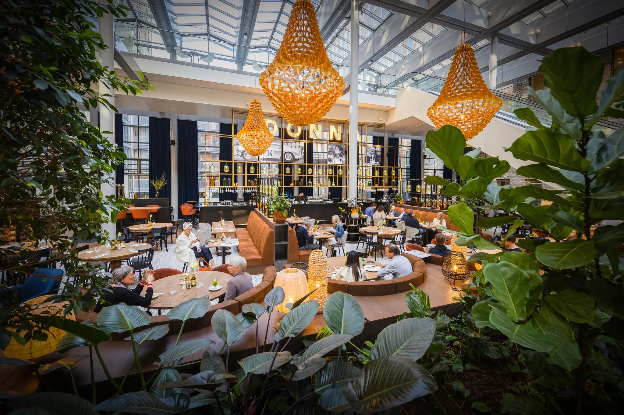 Modern restaurant aan de Prinses Beatrixlaan met glazen dak, hanglampen en gasten aan stijlvolle zitplaatsen tussen groen.